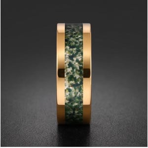 May include: A gold-toned metal ring featuring a wide, flat band with a central inlay of mossy green and white speckled stone material. The polished metal edges frame the textured, organic-looking inlay, creating a distinct contrast between the smooth, reflective gold surface and the earthy, mottled green center.