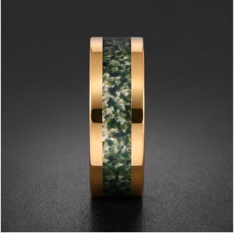 May include: A gold-toned metal ring featuring a wide, flat band with a central inlay of mossy green and white speckled stone material. The polished metal edges frame the textured, organic-looking inlay, creating a distinct contrast between the smooth, reflective gold surface and the earthy, mottled green center.