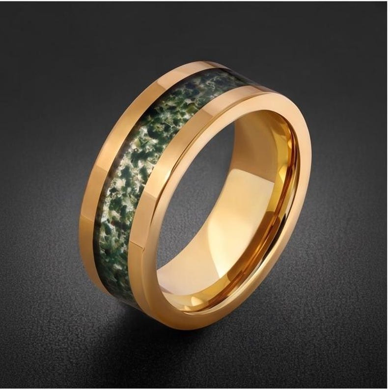 May include: A gold-toned band ring featuring a wide, central inlay of moss agate stone. The stone inlay displays a mottled pattern of deep forest green, white, and dark grey inclusions, set within a polished, reflective gold-colored metal band.
