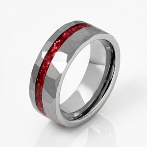 May include: A silver-toned ring featuring a hammered, faceted exterior surface and a central inlay of crushed, vibrant red material that runs horizontally around the entire circumference of the band, creating a distinct contrast in texture and colour.