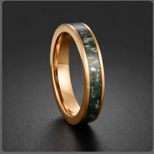 May include: A polished rose gold-toned ring featuring a central inlay of dark green and white mottled stone. The band has a smooth, reflective metallic finish on the edges, framing the textured, organic appearance of the stone inlay.