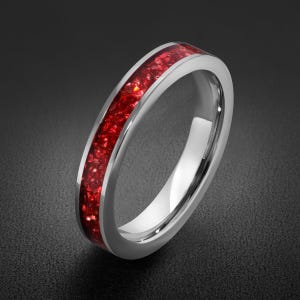 May include: A silver-toned metal ring featuring a central inlay of crushed, vibrant red stone fragments. The band has a smooth, polished finish on the edges, contrasting with the textured, sparkling appearance of the deep red inlay material.