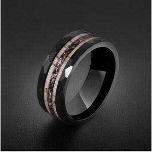 Pu&ograve; includere: Un anello nero con una superficie esterna sfaccettata e un intarsio centrale caratterizzato da una fascia di colore chiaro con una striscia centrale scura, testurizzata e dall'aspetto metallico. L'anello ha una finitura interna liscia.