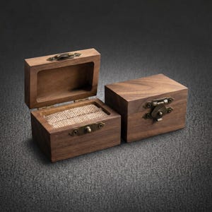 May include: Two rectangular wooden boxes made of dark brown walnut wood. One box is open, revealing a textured beige burlap insert for holding rings, while the other box is closed. Both feature antique brass-colored metal latches and hinges.