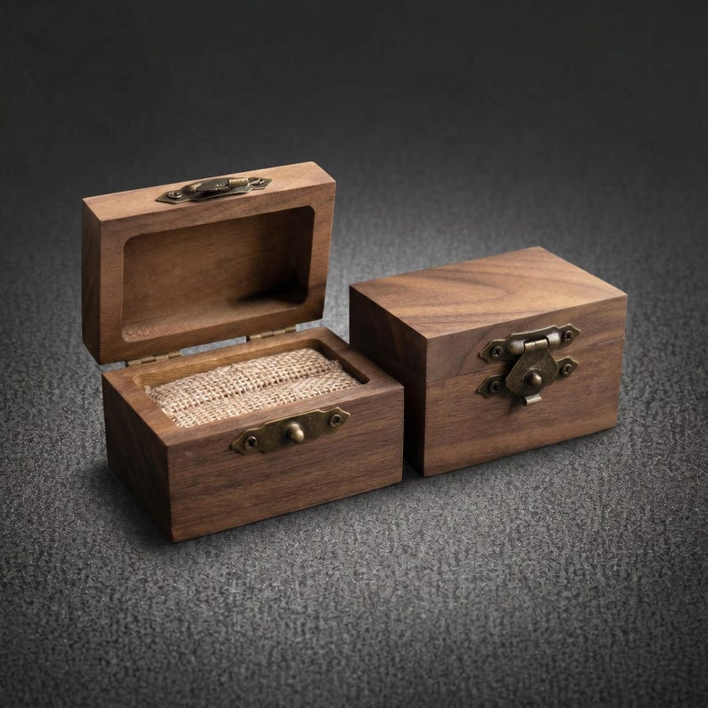 May include: Two rectangular wooden boxes made of dark brown walnut wood. One box is open, revealing a textured beige burlap insert for holding rings, while the other box is closed. Both feature antique brass-colored metal latches and hinges.