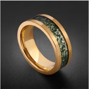 May include: A gold-toned band ring featuring a central inlay of mossy green and white mottled stone. The ring has a polished, reflective metallic finish on the edges and a smooth, rounded interior surface, showcasing a distinct contrast between the metal.