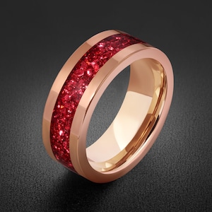 May include: A rose gold-toned ring featuring a wide, polished band with a central inlay of crushed, deep red crystalline material. The ring has a smooth, reflective metallic finish on the edges and a hollow centre, showcasing a clean, circular design.