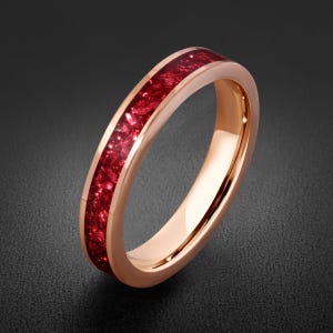 May include: A rose gold-toned ring featuring a central inlay of crushed, deep red crystalline material. The band has a smooth, polished metallic finish with a rounded profile, showcasing a vibrant contrast between the metal and the red inlay.