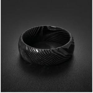 May include: A black ring featuring a distinct, wavy, layered pattern across its entire outer surface. The band has a wide, flat profile with a dark, matte finish that highlights the intricate, flowing lines etched into the metal material.