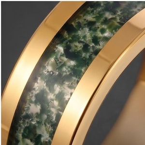 May include: A gold-toned ring featuring a central inlay of moss agate stone. The band has polished, smooth gold-colored edges that frame the textured, mottled green and white stone inlay, creating a distinct contrast between the metal and stone.