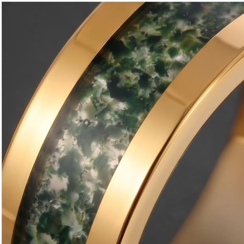 May include: A gold-toned ring featuring a central inlay of moss agate stone. The band has polished, smooth gold-colored edges that frame the textured, mottled green and white stone inlay, creating a distinct contrast between the metal and stone.