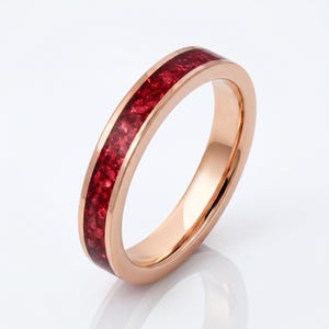 May include: A rose gold band ring featuring a central inlay of crushed deep red stones. The metal surface is smooth and polished, creating a reflective finish that contrasts with the textured, granular appearance of the vibrant red inlay material.