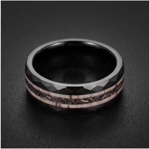 Pu&ograve; includere: Un anello nero con una superficie esterna sfaccettata e un inserto centrale caratterizzato da un motivo testurizzato marrone scuro e argento metallizzato. La fascia ha una finitura lucida e un profilo circolare ampio e distinto.