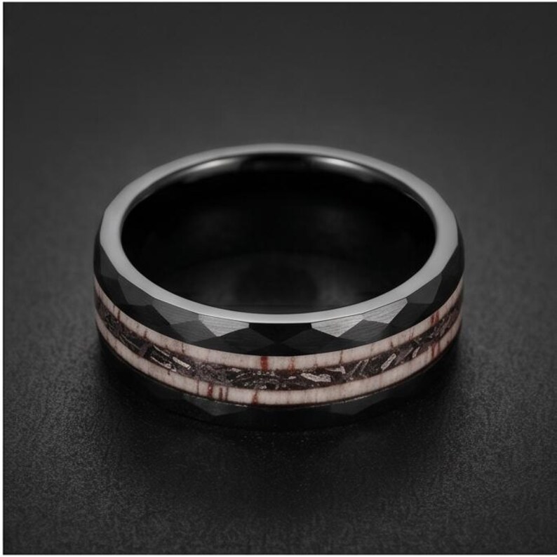 Pu&ograve; includere: Un anello nero con una superficie esterna sfaccettata e un inserto centrale caratterizzato da un motivo testurizzato marrone scuro e argento metallizzato. La fascia ha una finitura lucida e un profilo circolare ampio e distinto.