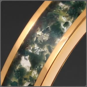 May include: A curved gold-toned ring featuring a central inlay of translucent resin with swirling dark green, teal, and mossy patterns interspersed with reflective silver-colored metallic flakes throughout the entire length of the band.