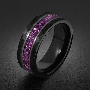 Op de afbeelding: Een zwarte ring met een gefacetteerd buitenoppervlak en een centrale inleg van vermalen paarse steen. De ring heeft een gladde binnenafwerking en een gepolijst donker metallic uiterlijk, wat een contrast vormt tussen het donkere metaal en de paarse inleg.