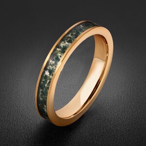 May include: A rose gold-toned ring featuring a smooth, polished metal band with a central inlay of moss agate. The stone inlay displays a dark green, mottled, and forest-like pattern set flush within the metallic channel of the ring band.