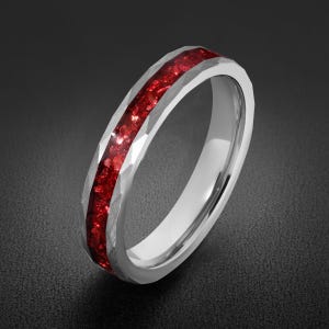 Op de afbeelding: Een zilverkleurige ring met een gefacetteerd, gehamerd buitenoppervlak en een centrale inleg van vermalen, dieprood materiaal. De ring heeft een gepolijste binnenkant en een opvallende, getextureerde metalen rand rondom de rode inleg.