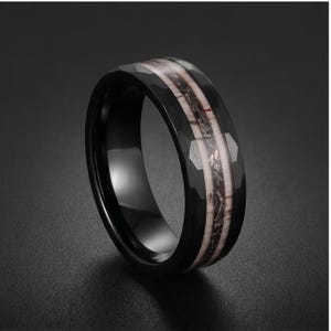 Pu&ograve; includere: Un anello nero con superficie esterna sfaccettata e un inserto centrale caratterizzato da un motivo testurizzato nei toni del chiaro e dello scuro. La fascia presenta una finitura interna liscia e lucida con una struttura ampia e robusta.