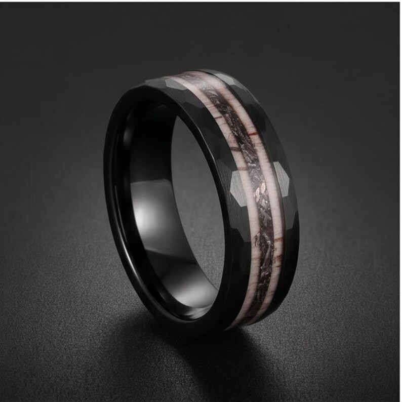 Pu&ograve; includere: Un anello nero con superficie esterna sfaccettata e un inserto centrale caratterizzato da un motivo testurizzato nei toni del chiaro e dello scuro. La fascia presenta una finitura interna liscia e lucida con una struttura ampia e robusta.