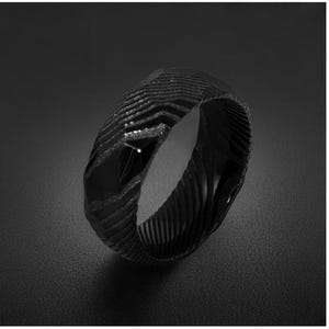 May include: A black ring featuring a textured, multi-faceted surface with a repeating chevron pattern. The band has a dark, matte finish with alternating smooth and granular sections that create a geometric, angular appearance around the circumference.