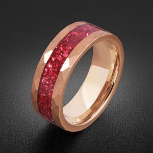 May include: A rose gold-toned ring featuring a hammered exterior finish and a central inlay of crushed red stone fragments. The band has a smooth, polished interior surface and a wide, flat profile with a distinct textured metallic appearance.