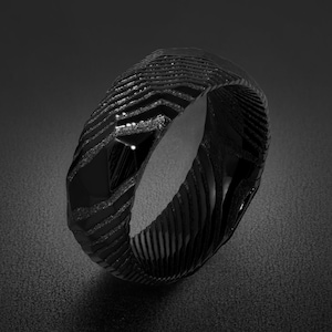 May include: A black ring featuring a textured, layered surface with a series of repeating, angular, chevron-like grooves and polished, faceted sections that create a high-contrast appearance across the entire circumference of the band.