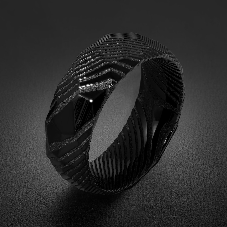 May include: A black ring featuring a textured, layered surface with a series of repeating, angular, chevron-like grooves and polished, faceted sections that create a high-contrast appearance across the entire circumference of the band.