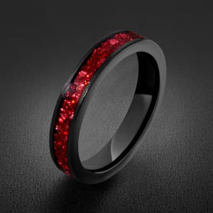 Puede incluir: Un anillo negro con una incrustación central de fragmentos de piedra roja triturada. La banda tiene un acabado liso y pulido con un perfil redondeado, donde la incrustación roja vibrante crea un contraste texturizado sobre el metal negro.