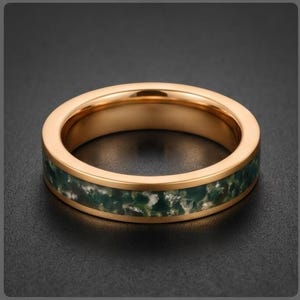 May include: A rose gold-toned band ring featuring a central inlay of dark green and white mottled stone. The ring has a smooth, polished metallic finish on the edges and a textured, organic appearance within the inlaid section of the band.
