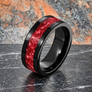 May include: A black ring featuring a wide, polished band with a central inlay of crushed, vibrant red material. The surface of the ring is smooth and reflective, contrasting with the textured, granular appearance of the red inlay running around the centre.