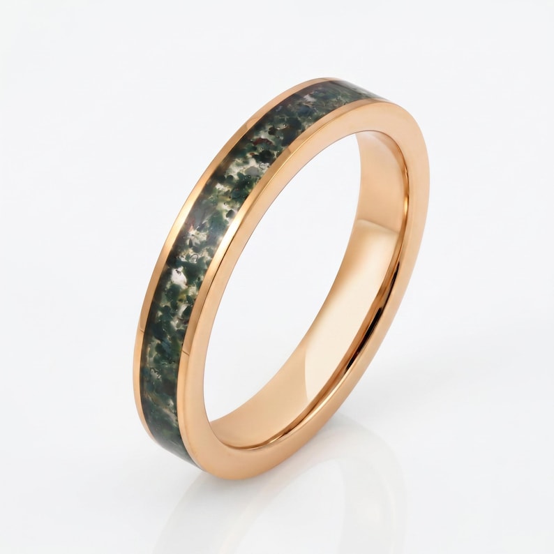 May include: A rose gold-toned ring featuring a central inlay of moss agate stone. The band has a smooth, polished metallic finish on the edges, while the center channel is filled with a textured, dark green and white speckled mineral pattern.