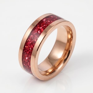 May include: A rose gold-toned ring featuring a wide, polished metal band with a central inlay of crushed, deep red, sparkling material that runs continuously around the circumference of the ring, creating a textured and reflective appearance.