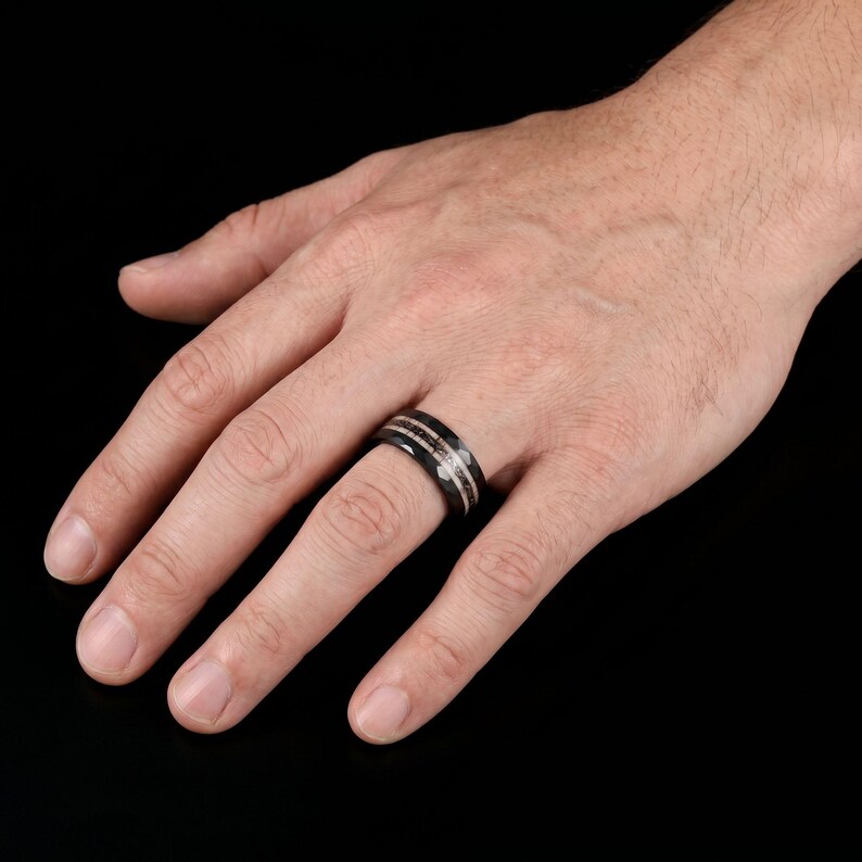 Pu&ograve; includere: Un anello nero caratterizzato da un inserto centrale con un aspetto testurizzato, metallico e maculato marrone scuro. La fascia presenta una finitura liscia e lucida sui bordi esterni, creando un netto contrasto con l'inserto centrale dettagliato.