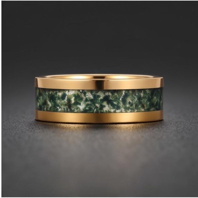May include: A gold-toned metal ring featuring a wide, flat band with a central inlay of moss agate. The stone inlay displays a mottled, organic pattern of deep forest green and translucent white inclusions, framed by polished gold-colored edges.