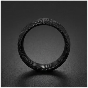 May include: A black ring featuring a textured surface with a repeating, diagonal, wave-like pattern that wraps around the entire band. The metal has a matte finish and a dark, monochromatic appearance, showcasing a consistent, intricate, and grooved design.