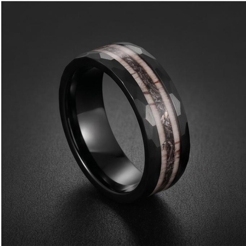Pu&ograve; includere: Un anello nero con superficie esterna sfaccettata e un inserto centrale caratterizzato da un motivo testurizzato color marrone scuro e crema. La fascia presenta una finitura interna liscia e un aspetto metallico scuro sui bordi.
