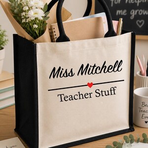 May include: A canvas tote bag with black trim and handles, personalized with "Miss Mitchell" and "Teacher Stuff." The bag contains a bouquet of white flowers, notebooks, and a pen. A mug that says "Best Teacher Ever" is also visible.