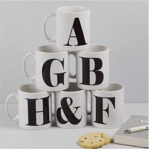 Puede incluir: Tazas de cerámica blancas con letras mayúsculas negras A, G, B, H, &, y F. Las tazas están apiladas en forma de pirámide. Un pequeño cuaderno, bolígrafo y galletas están al lado. Las tazas tienen un diseño simple y moderno.