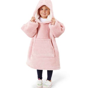 Puede incluir: Una manta de forro polar rosa con capucha y un gran bolsillo en la parte delantera. La manta la lleva puesta un ni&ntilde;o y tiene un forro de sherpa blanco alrededor de la capucha.
