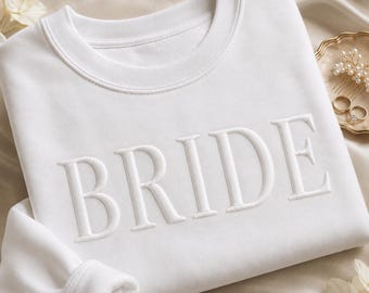 May include: A white crewneck sweatshirt featuring the word BRIDE embroidered in matching white thread across the chest. The garment has a ribbed collar and long sleeves, presented with a gold hair accessory and two rings nearby.