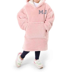 Puede incluir: Una manta de forro polar rosa con capucha y un gran bolsillo en la parte delantera. La manta tiene las iniciales "MJ" bordadas en gris en la parte delantera.