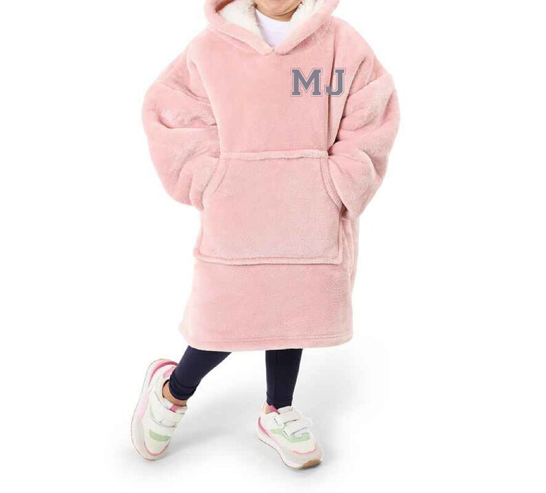 Puede incluir: Una manta de forro polar rosa con capucha y un gran bolsillo en la parte delantera. La manta tiene las iniciales "MJ" bordadas en gris en la parte delantera.