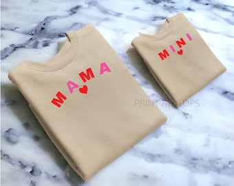 May include: Two tan sweatshirts with the words "MAMA" and "MINI" in pink and red letters with a pink heart on each.