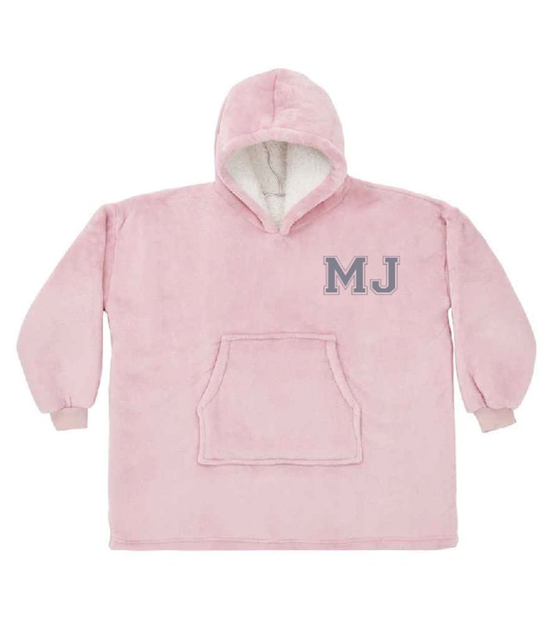 Puede incluir: Una manta de forro polar rosa con capucha y un gran bolsillo en la parte delantera. La manta tiene las iniciales "MJ" bordadas en gris en la parte delantera.