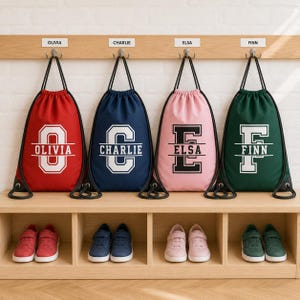 May include: Four drawstring bags in red, navy blue, pink, and dark green, each featuring a large white letter and a name printed across it. The bags hang from silver hooks on a wooden rack above four pairs of matching colored sneakers.