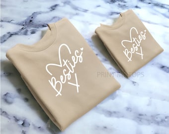 May include: Two tan sweatshirts with the text "Besties" in white lettering with a heart design.