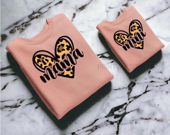 May include: Two pink sweatshirts with a heart design. The larger sweatshirt has the word "mama" in black text with a leopard print heart. The smaller sweatshirt has the word "mini" in black text with a leopard print heart.