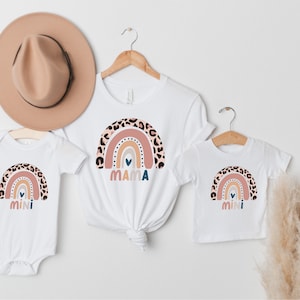 Pode incluir: Um conjunto de três t-shirts brancas com um design de arco-íris com estampa de leopardo, um coração e as palavras "mama" e "mini".