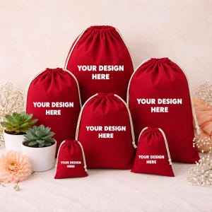 Könnte beinhalten: Eine Sammlung von fünf roten Kordelzugbeuteln in verschiedenen Größen, die jeweils mit dem weißen Text YOUR DESIGN HERE versehen sind. Die Beutel bestehen aus Stoff und haben cremefarbene Kordeln zum Verschließen des Inhalts.